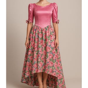 May include: A pink satin dress with a floral print skirt. The bodice has a sweetheart neckline and short, puffed sleeves with bow details. The skirt features a high-low hemline, showcasing a vibrant rose pattern in shades of pink and green.