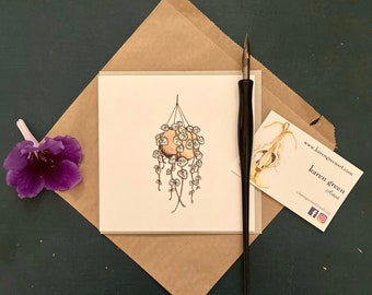 May include: A hand-drawn illustration of a hanging plant with round, light green leaves. The plant is in a brown pot and is hanging from a string. The illustration is on a white card with a brown envelope and a business card.