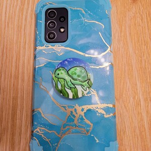 May include: A blue phone case with a marble pattern and gold accents. A pop socket with a green turtle design is attached to the back of the case.