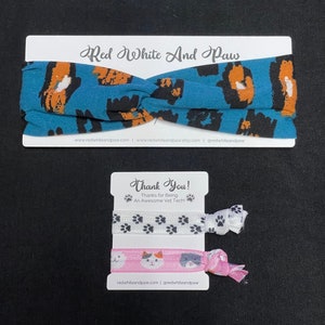May include: A thank you card with a blue and orange patterned headband and two hair scrunchies. The scrunchies have paw prints and a cat design. The card says "Thank You! Thanks for Being An Awesome Vet Tech!" and includes the website address redwhiteandpaw.com.