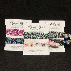 Op de afbeelding: Drie sets met kleurrijke haarbandjes met hondenthema's. Elke set bevat drie haarbandjes met verschillende ontwerpen. De haarbandjes zijn verpakt op witte kaartjes met de tekst "Bedankt! Bedankt dat je een geweldige dierenartsassistent bent!" en contactgegevens van de verkoper.