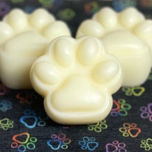 May include: Three white paw-shaped wax melts on a black background with colorful paw prints.