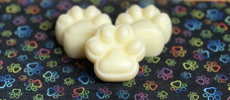 May include: Three white paw-shaped wax melts on a black background with colorful paw prints.