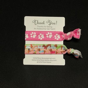 Op de afbeelding: Twee kleurrijke haarbandjes met pootprintontwerpen. Het roze haarbandje heeft witte pootprints en het andere haarbandje heeft een veelkleurig patroon van honden. De kaart zegt "Bedankt! Bedankt dat je een geweldige dierenarts bent!" en bevat contactgegevens van de verkoper.