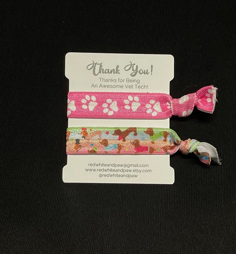 Op de afbeelding: Twee kleurrijke haarbandjes met pootprintontwerpen. Het roze haarbandje heeft witte pootprints en het andere haarbandje heeft een veelkleurig patroon van honden. De kaart zegt "Bedankt! Bedankt dat je een geweldige dierenarts bent!" en bevat contactgegevens van de verkoper.