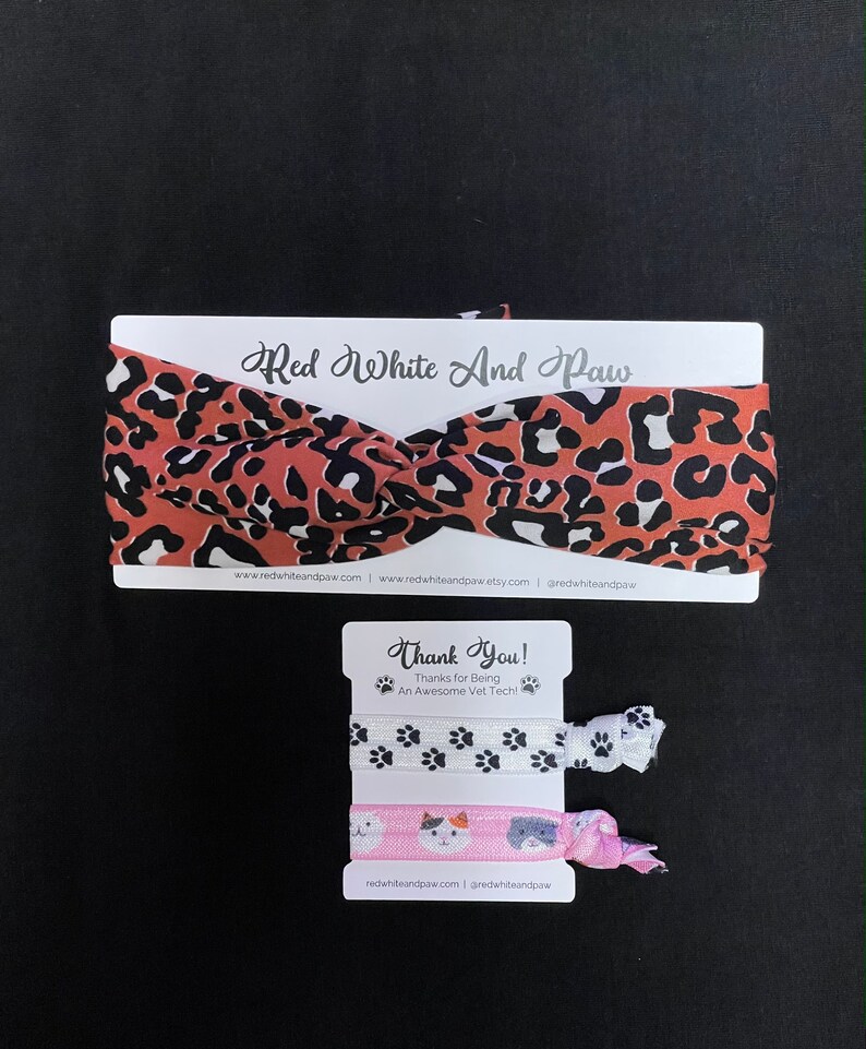 May include: A set of two hair scrunchies with a leopard print headband. The scrunchies feature paw prints and a cat design. The headband is red with black and white spots. The packaging includes the text "Red White And Paw" and "Thank You! Thanks for Being An Awesome Vet Tech!"