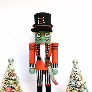 May include: A hand-painted wooden nutcracker with a green zombie face, red and black striped trousers, and a black top hat. The nutcracker is holding a silver knife and standing on a red base. There are two small artificial Christmas trees with colourful baubles on either side of the nutcracker.