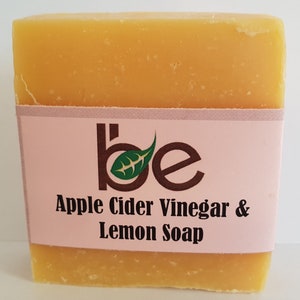 Peut inclure: Un pain de savon rectangulaire de couleur orange clair. Une étiquette rose entoure le savon, affichant le texte "be" avec un motif de feuille et les mots "Apple Cider Vinegar & Lemon Soap" en noir. Le savon semble fait à la main.