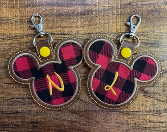 May include: Two Mickey Mouse-shaped keychains with a red and black plaid pattern. Each keychain has a brown border and a yellow initial. The keychains have a silver clasp and a brown leather strap.