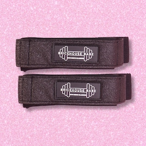 Op de afbeelding: Een paar zwarte gewichthefslingers met een glinsterende afwerking. De slingers hebben een wit logo met de tekst "Brickhouse Babe Gym Meets Glamour".