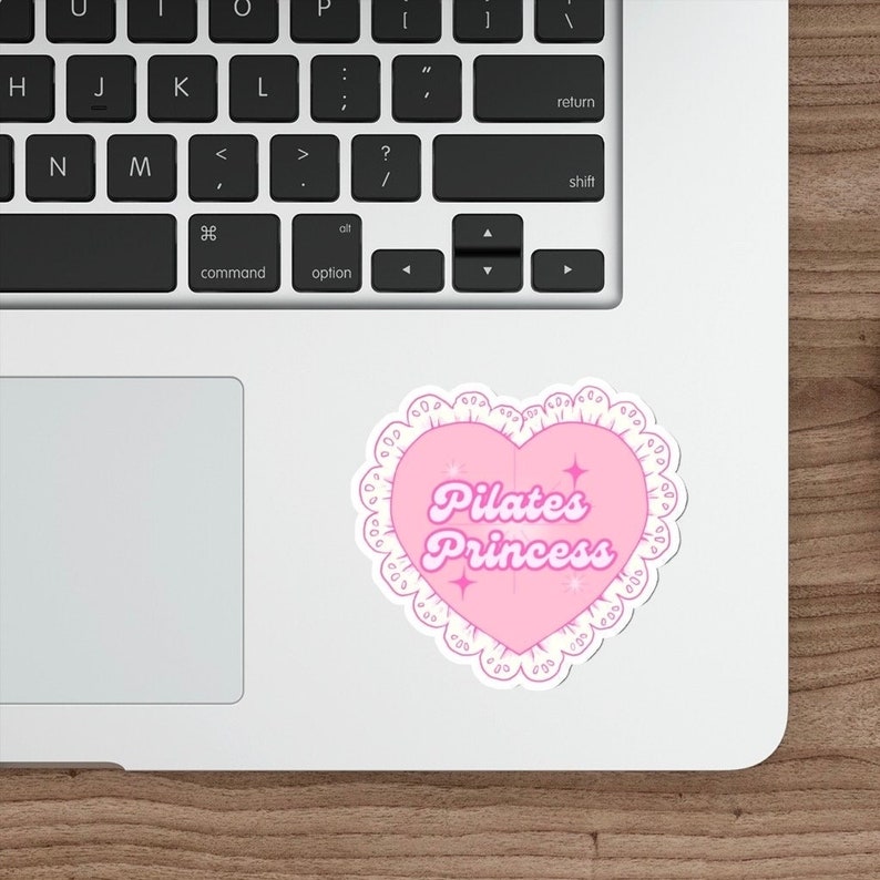 May include: A pink heart-shaped sticker with the text "Pilates Princess" in a cursive font. The sticker has a white lace-like border and small star accents. The sticker is placed on a laptop.