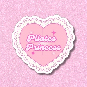 Pode incluir: Um autocolante rosa em forma de coração com a frase "Pilates Princess" em fonte retro. O coração é contornado por uma borda branca recortada e adornado com detalhes de estrelas, sobre um fundo rosa brilhante. Para entusiastas de pilates.
