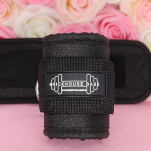 May include: Black weightlifting wrist wraps with the text "BRICKHOUSE BABE GYM MEETS GLAMOUR" on a rectangular patch. The wraps are made of a textured material and are set against a backdrop of pink and white roses.