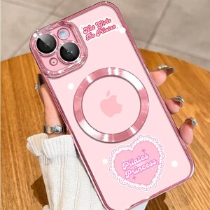 May include: Pink phone case with a clear camera lens protector and a magnetic ring. The case features the text "Hot Girls Do Pilates" and a heart-shaped sticker that says "Pilates Princess".