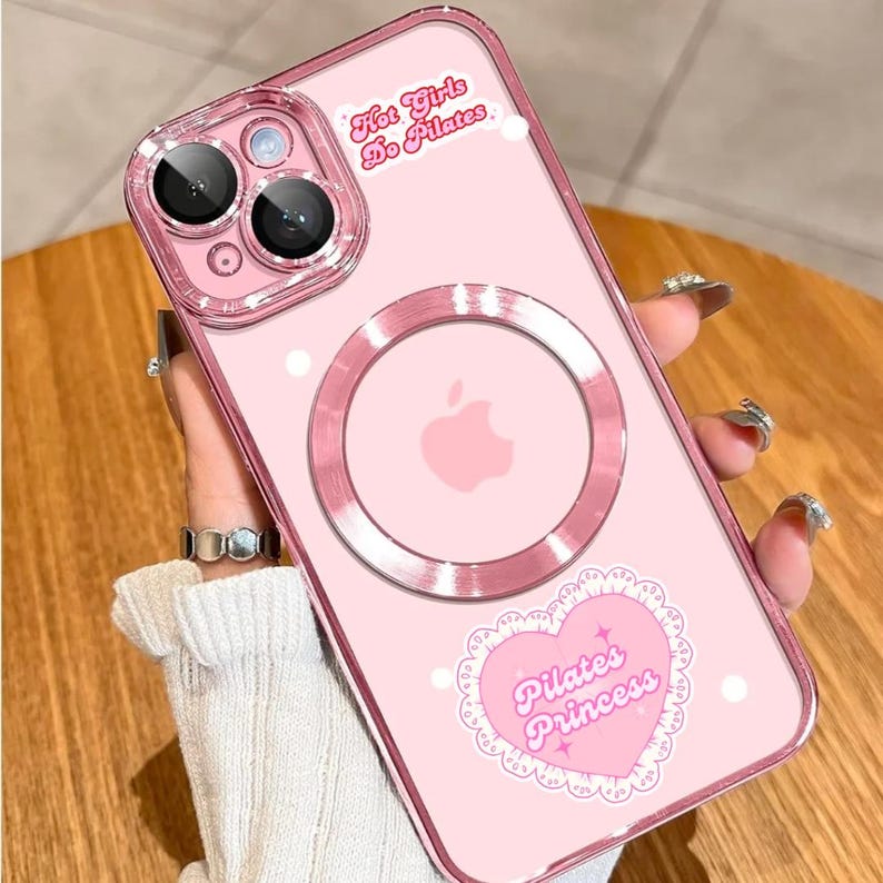 May include: Pink phone case with a clear camera lens protector and a magnetic ring. The case features the text "Hot Girls Do Pilates" and a heart-shaped sticker that says "Pilates Princess".