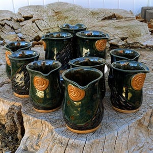 May include: A collection of handmade ceramic pitchers with a dark green glaze. Each pitcher features a unique design, including spiral and textured patterns, and a light brown base. The pitchers are arranged on a weathered wooden surface.