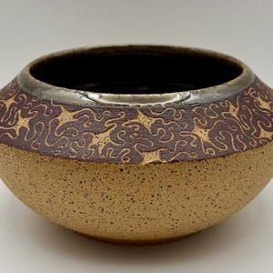 May include: A round ceramic bowl with a speckled tan base and a dark brown upper band featuring an intricate, swirling, light-colored pattern. The interior of the bowl is dark, and the rim has a metallic, reflective finish with a smooth texture.