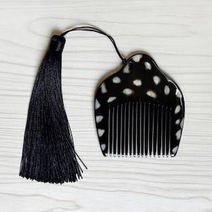 May include: A black comb with white speckled detailing and a long black tassel. The comb features a curved top and a series of thin, parallel teeth. The tassel is made of many strands of black thread.