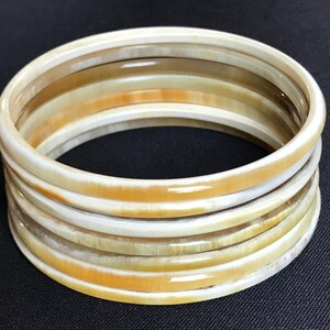 May include: A set of eight ivory-colored bangles with a natural wood grain pattern. The bangles are stacked on top of each other, creating a layered effect.
