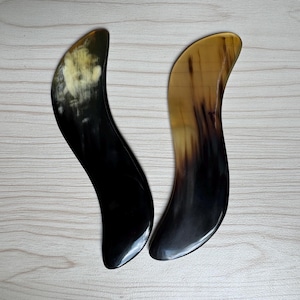 Puede incluir: Dos herramientas curvas, con forma de cuerno, con un acabado liso y pulido. Una es principalmente negra con un área más clara y moteada en la parte superior, mientras que la otra pasa de un marrón claro a un marrón más oscuro. Probablemente se utilizan para masajes o cuidado de la piel.