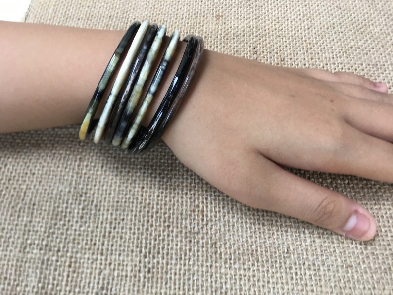 May include: A set of black and white patterned bangles. The bangles are made of a hard, smooth material and are stacked on a person's wrist.
