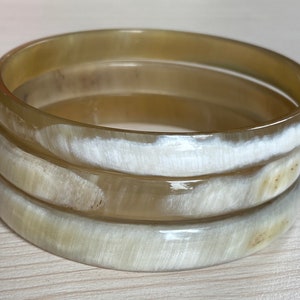 Peut inclure: Trois bracelets en corne empilés de couleur beige clair avec un motif de rayures blanches.