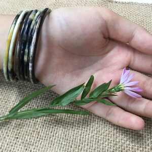 May include: A set of five brown, black, and yellow bangles worn on a wrist. The bangles are made of a patterned material. A small purple flower with green leaves is resting on the palm of a hand.