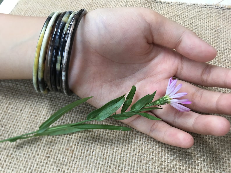 May include: A set of five brown, black, and yellow bangles worn on a wrist. The bangles are made of a patterned material. A small purple flower with green leaves is resting on the palm of a hand.