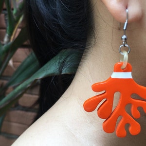 Puede incluir: Pendientes colgantes de color naranja y blanco con forma de coral. Los pendientes están hechos de plástico y tienen una franja blanca en la parte superior.