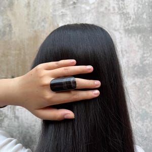 Puede incluir: Un anillo grande, rectangular, negro y marrón con un acabado pulido. El anillo se lleva en un dedo, con pelo largo y oscuro en el fondo. También se ve una pulsera plateada.