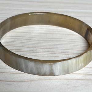 May include: A round, polished bracelet with a natural horn appearance. The bracelet has a light brown and cream color with a smooth, glossy finish. The bangle is a simple, elegant accessory.