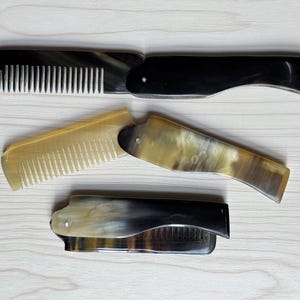 May include: Three folding pocket combs made of horn are displayed on a light wood surface. The combs are in shades of black, brown, and tan. The teeth of the combs are white. The combs are open and closed.