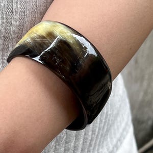 May include: A wide, glossy bangle bracelet in shades of brown, black, and yellow is worn on a person's arm. The bracelet has a smooth, curved design and appears to be made of a natural material. The bracelet is a fashion accessory.