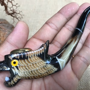 Puede incluir: Una pipa de tabaco artesanal con diseño de dragón. Presenta una combinación de colores negro y marrón con un acento metálico dorado. La cabeza del dragón está detallada con escamas, ojos amarillos y dientes blancos. El tallo es curvo y pulido.