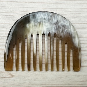 Puede incluir: Un peine semicircular con apariencia de cuerno natural. El peine presenta una serie de dientes espaciados uniformemente y un acabado liso y pulido. Los colores varían del marrón claro al marrón oscuro, con zonas blancas y crema.