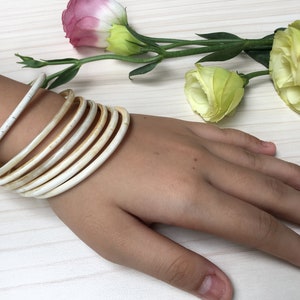 May include: A set of white bangle bracelets stacked on a person's wrist. The bracelets are made of a smooth, ivory-colored material.