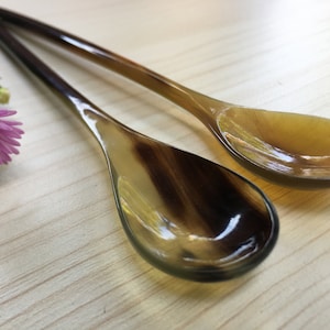 May include: Two brown and black horn spoons on a light wood surface. The spoons are oval shaped with a rounded end.