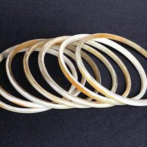 May include: A set of seven ivory-colored bangles made of horn. The bangles are stacked on top of each other, creating a layered effect.