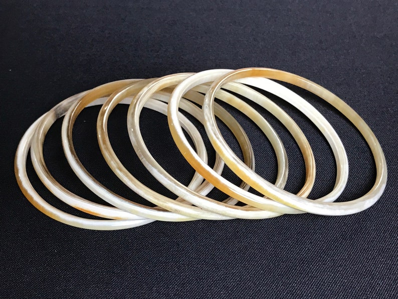May include: A set of seven ivory-colored bangles made of horn. The bangles are stacked on top of each other, creating a layered effect.