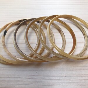 May include: A set of six horn bangles in a light brown color. The bangles are stacked on top of each other and have a smooth, polished finish.