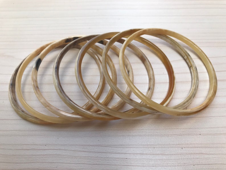 May include: A set of six horn bangles in a light brown color. The bangles are stacked on top of each other and have a smooth, polished finish.