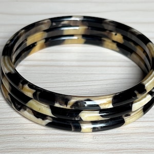 Op de afbeelding: Drie zwarte en crèmekleurige armbandjes met een patroon. De armbandjes zijn gemaakt van plastic en hebben een glanzende afwerking.