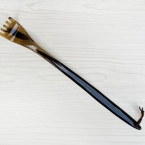 May include: A long, dark brown shoe horn with a forked end. The horn has a smooth, polished finish and a dark brown leather loop at the end. The shoe horn is made of a dark brown material.