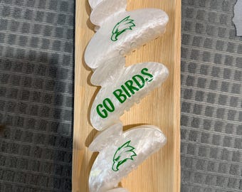 May include: Five pearlescent white hair claw clips, displayed on a wooden tray. The clips are inscribed with the words "EAGLES" and "GO BIRDS" in green, alongside a green eagle emblem. The tray is rectangular.