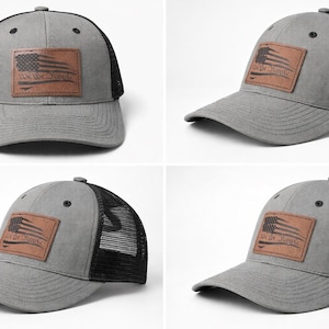 Op de afbeelding: Grijze truckerpet met een zwarte mesh achterkant en een gebogen klep. Een bruine leren patch op de voorkant toont een Amerikaans vlagontwerp met de tekst "We the People". De pet heeft een snapback sluiting.
