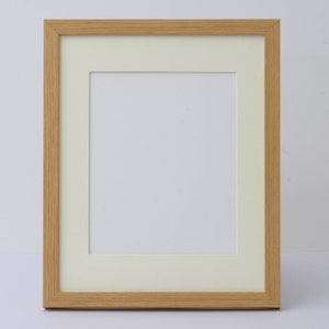May include: A rectangular picture frame with a light wood grain finish. The frame has a cream-colored mat and a white interior space for a photo or artwork. The frame is designed for displaying images.