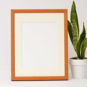 May include: A natural wood picture frame with a white mat and a blank white space inside. The frame is sitting on a white surface next to a potted snake plant.