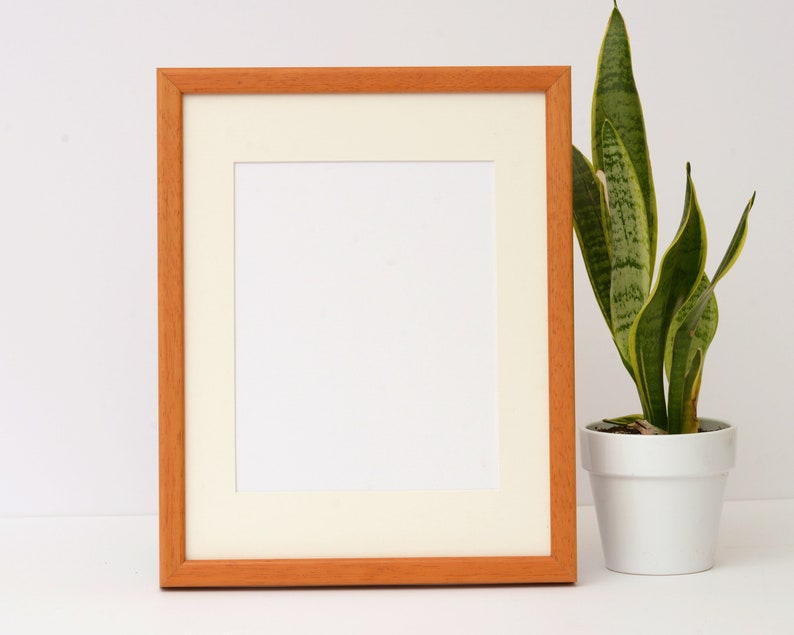 May include: A natural wood picture frame with a white mat and a blank white space inside. The frame is sitting on a white surface next to a potted snake plant.