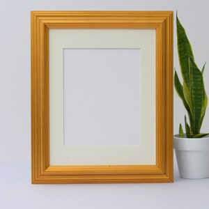 May include: A gold picture frame with a white mat and a blank white space inside. The frame is sitting on a white surface next to a potted plant with green leaves.