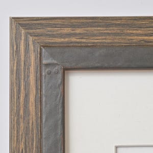 May include: Close-up of a wooden picture frame with a metal trim. The frame has a dark brown wood finish with a light brown grain pattern. The metal trim is a dark gray color and has a slightly distressed look. The frame is designed to hold a picture or artwork.